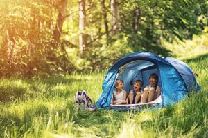 Little girl and brothers are camping in a tent in a sunny forest. Kids aged 5 and 9 are smiling happiliy sitting inside of blue tent. Sunny summer day. Slightly soft.